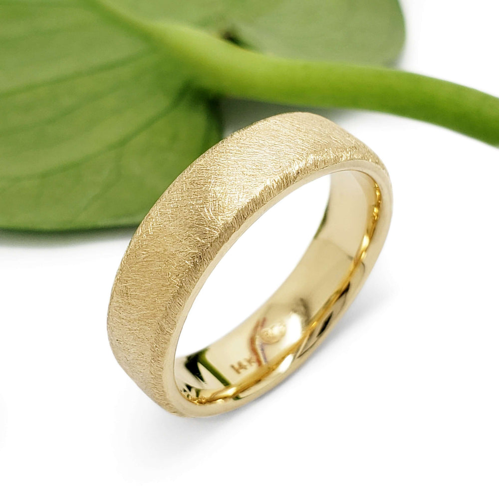 Flat 14kt yellow gold wedding ring with saw blade texture beside a green leaf