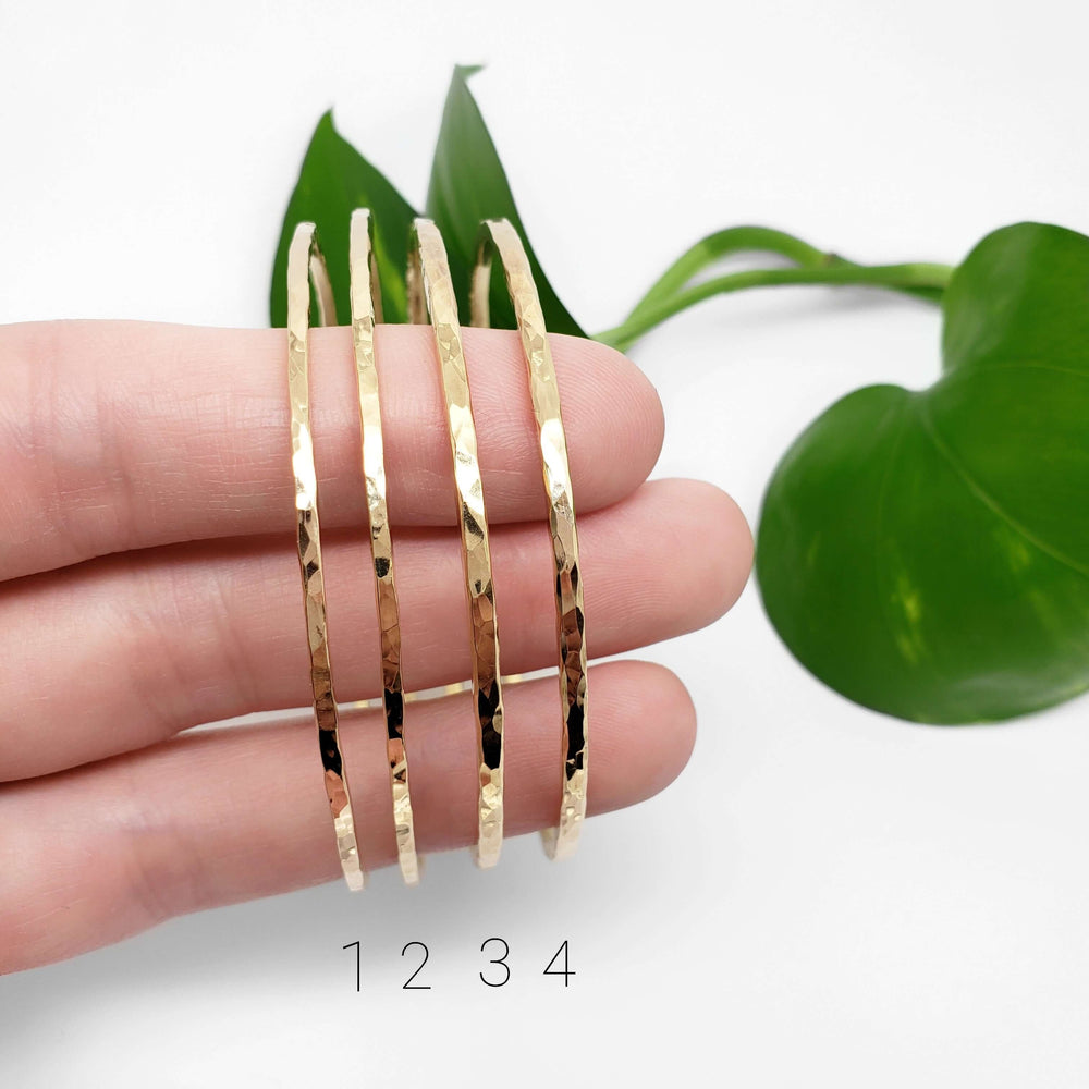 Four hammered 14kt yellow gold bangles displayed on fingers with green leaves