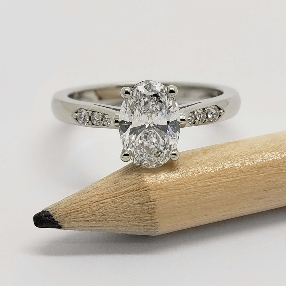Front view of oval lab diamond white gold engagement ring with pave band beside pencil