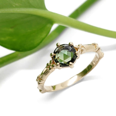 Green sapphire engagement ring in yellow gold with floral accents, shown with greenery