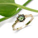 Green sapphire engagement ring in yellow gold with floral accents, shown with greenery