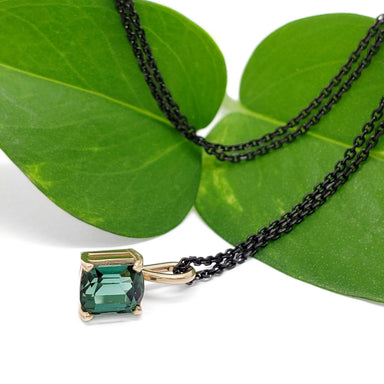 Barrel cut green tourmaline pendant in yellow gold with black chain, styled on green leaves