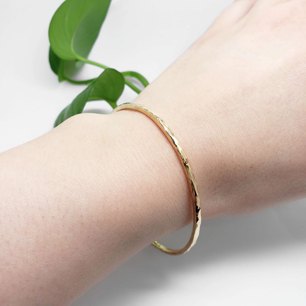 Single hammered 14kt yellow gold bangle worn on wrist with green leaves