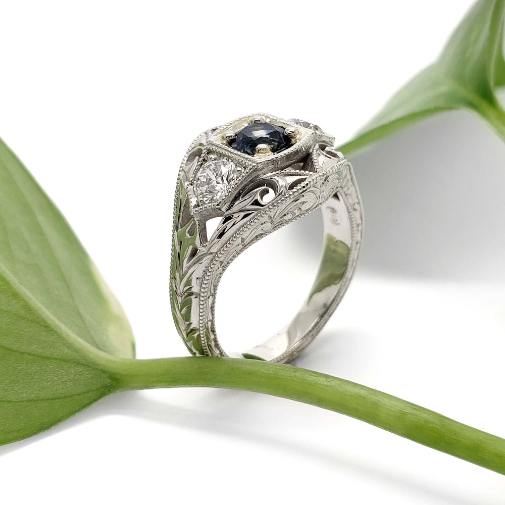 Hand-engraved white gold Montana sapphire and diamond ring beside a green leaf