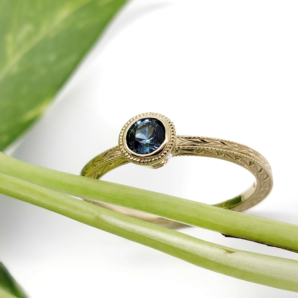 Hand-engraved yellow gold ring with teal Montana sapphire, shown with green leaves