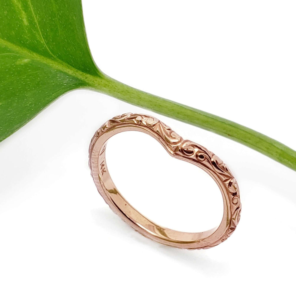 Hand engraved 14kt rose gold chevron wedding ring beside a green leaf