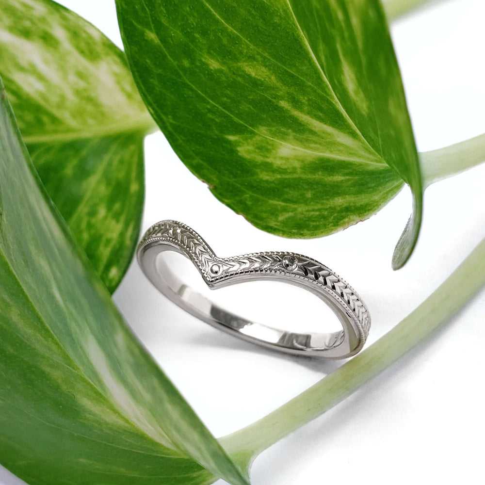 Hand engraved 14kt white gold chevron wedding ring displayed among green leaves