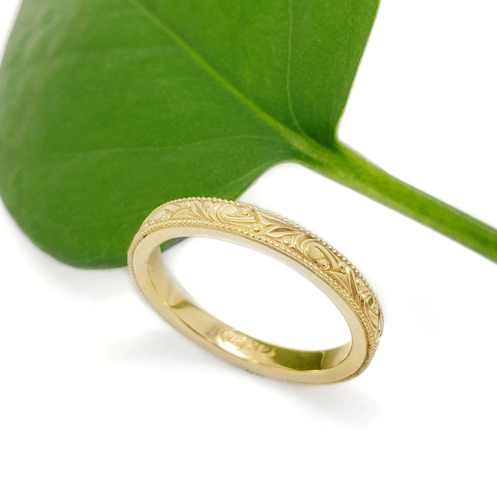 Hand engraved 14kt yellow gold wedding ring with millgrain, shown beside a green leaf
