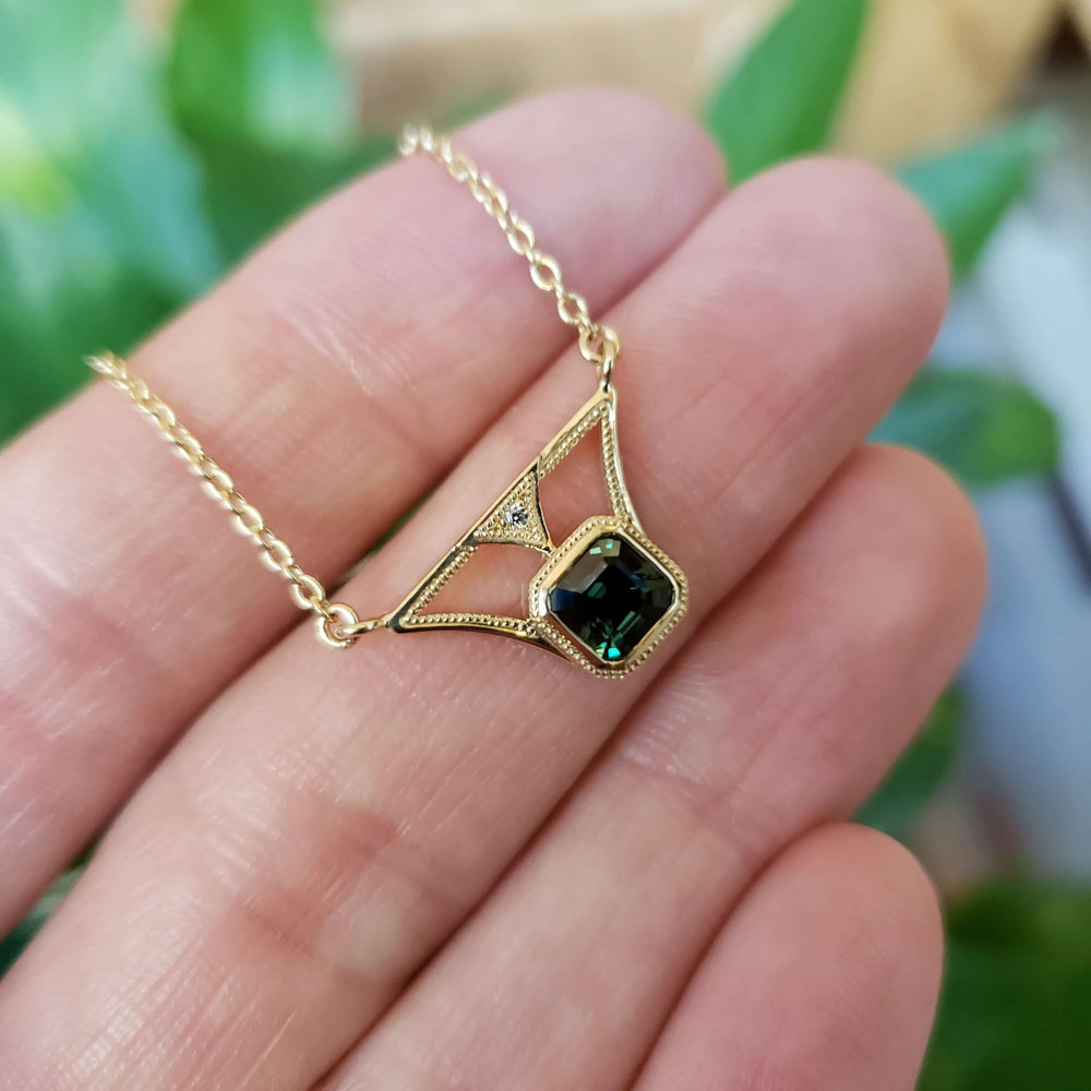 Hand holding yellow gold necklace with green asscher cut sapphire and diamond accent
