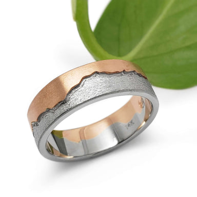 Handcrafted 14k white and rose gold mountain range ring beside a green leaf