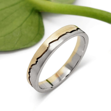 Handcrafted 14k white and yellow gold mountain range ring with brushed finish beside green leaf