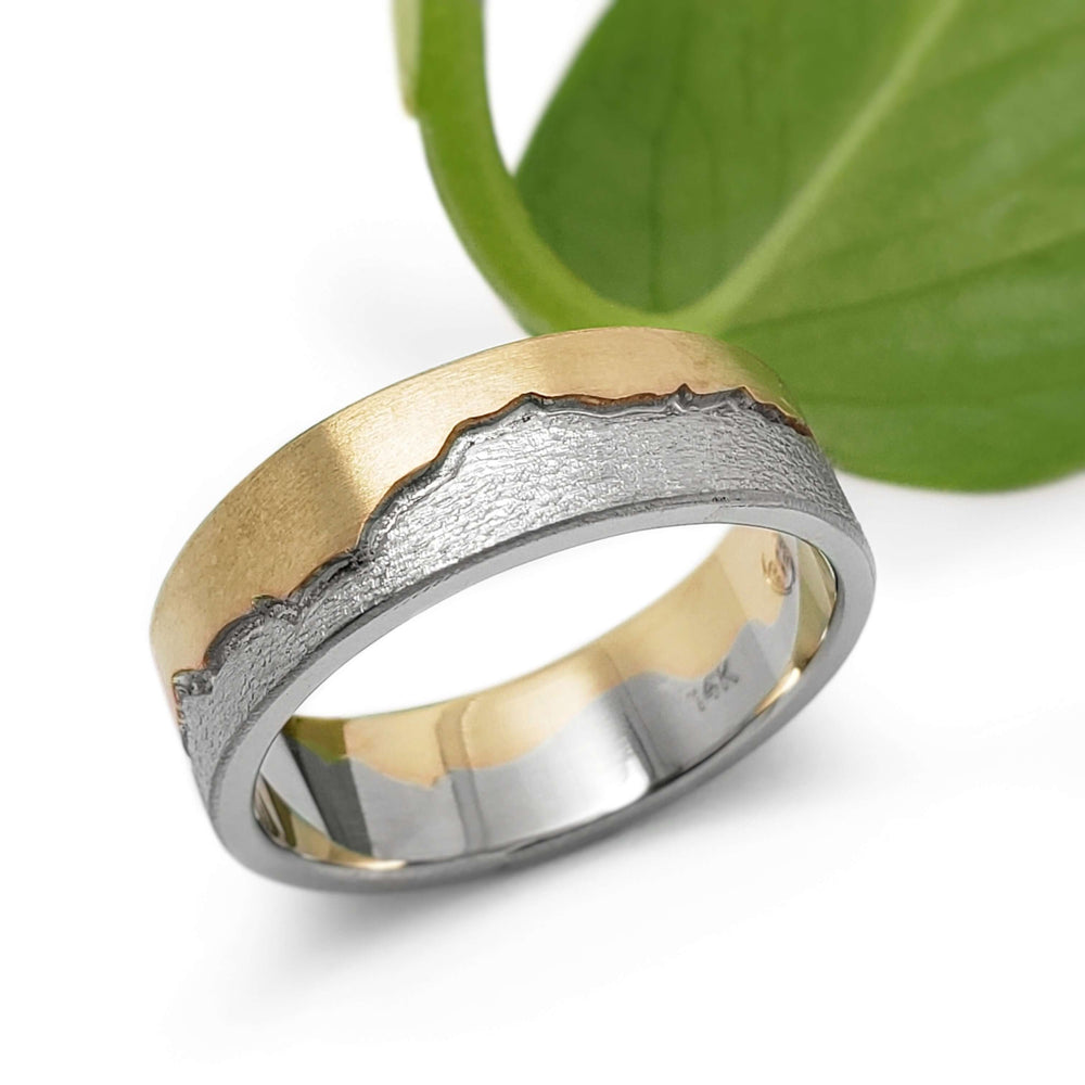 Handcrafted 14k white and yellow gold mountain range wedding band beside a green leaf