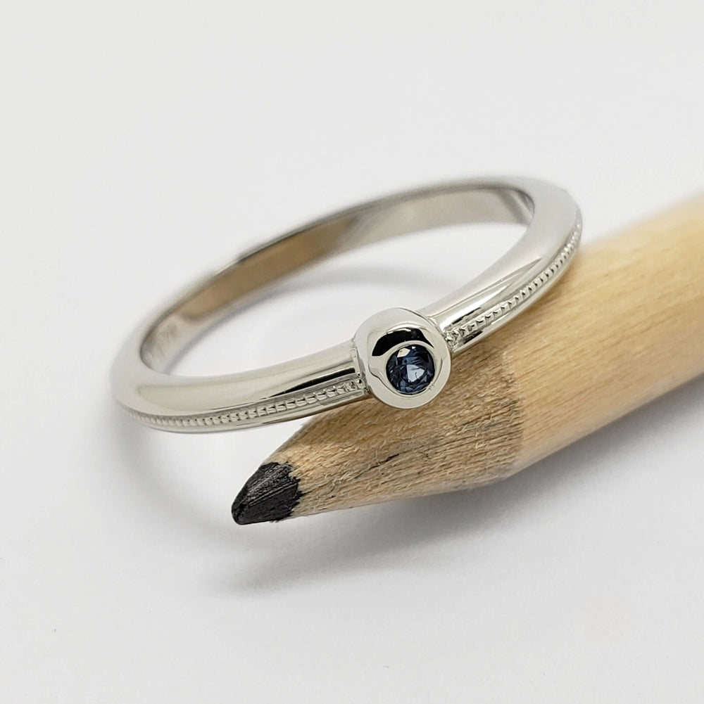 Handcrafted 14k white gold ring with blue sapphire, millgrain detail, beside pencil
