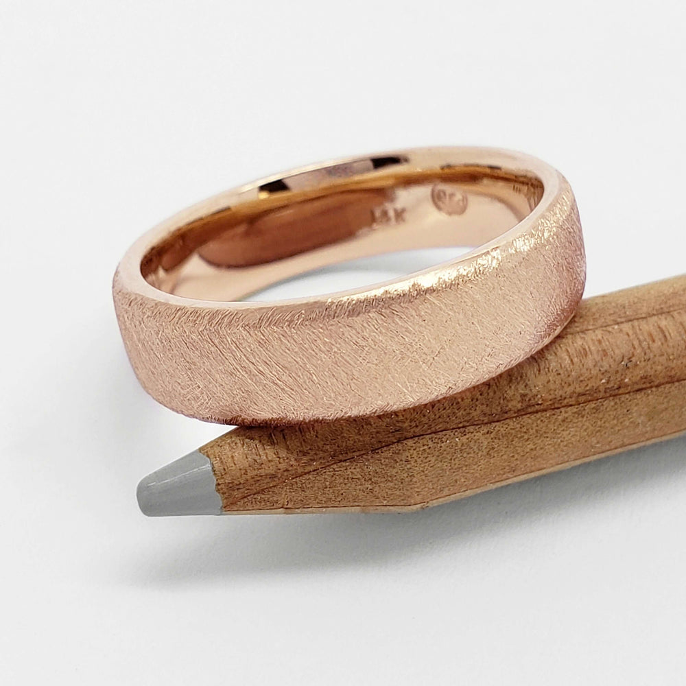 Handcrafted 14kt rose gold flat band with saw blade finish resting on a pencil