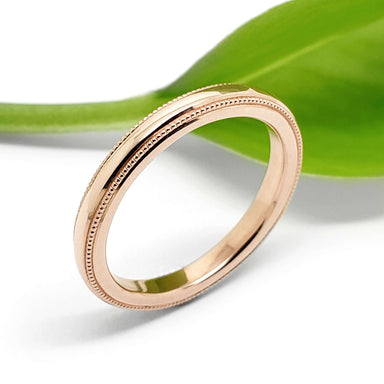 Handcrafted 14kt rose gold millgrain wedding ring beside a green leaf