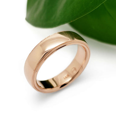 Handcrafted 14kt rose gold wedding ring with millgrain detail beside a green leaf