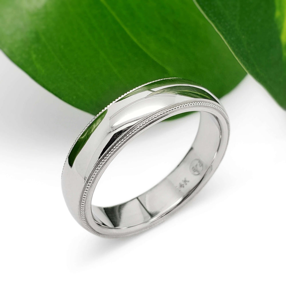Handcrafted 14kt white gold wedding ring with millgrain detail beside green leaf
