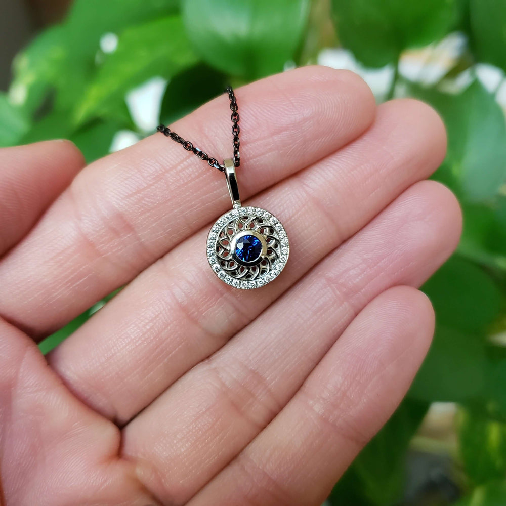 Hand holding 14kt white gold pendant with blue sapphire and diamond halo