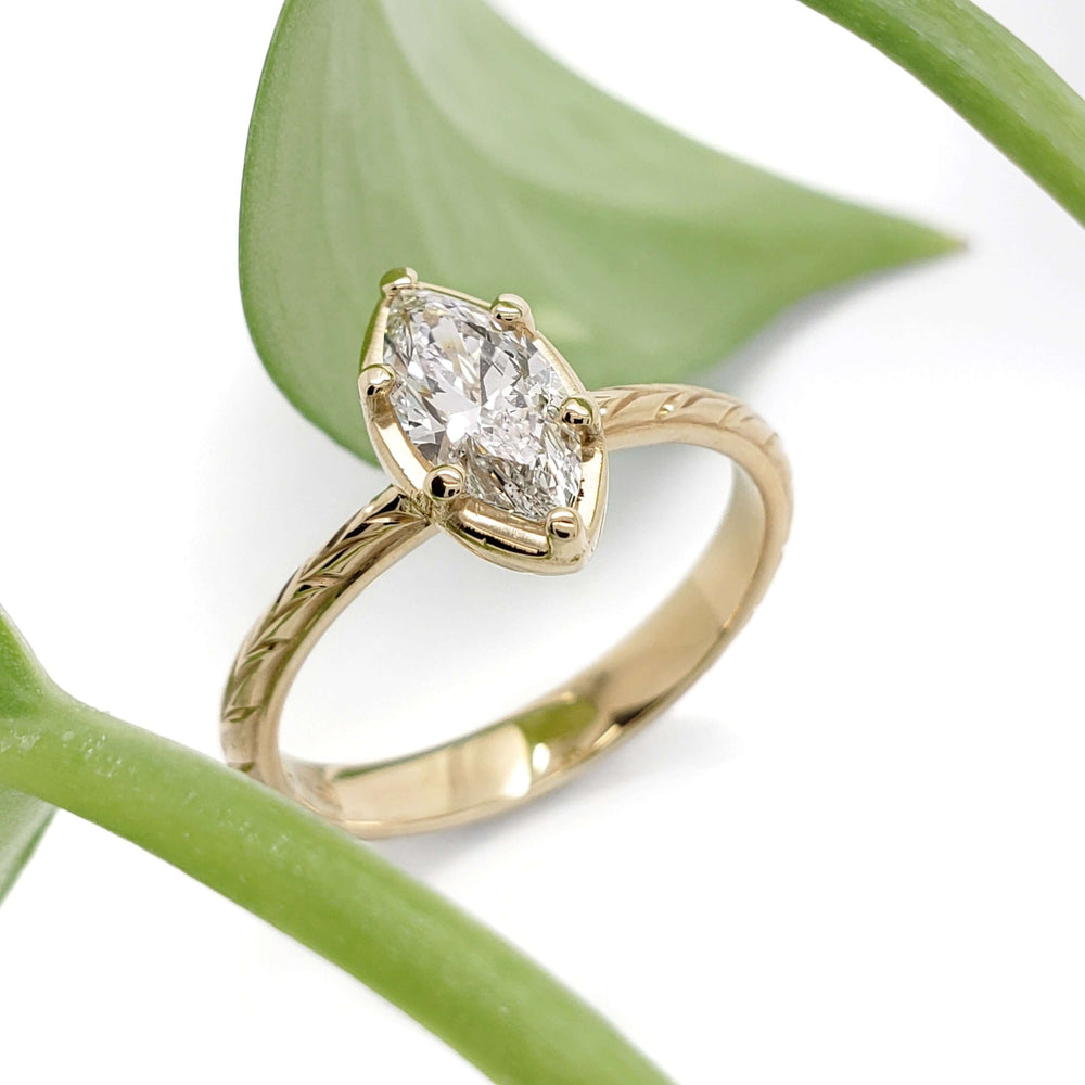 Handcrafted marquise lab diamond ring in 14kt yellow gold with engraved band, beside green leaf