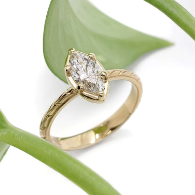 Handcrafted marquise lab diamond ring in 14kt yellow gold with engraved band, beside green leaf