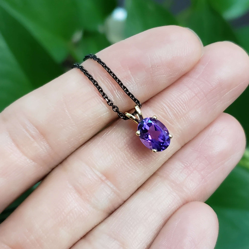 Hand holding oval amethyst pendant in 14kt yellow gold on black chain, with green background