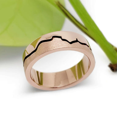 Handcrafted rose gold mountain range wedding band with blackened groove, beside green leaf