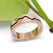 Handcrafted rose gold mountain range wedding band with blackened groove, beside green leaf