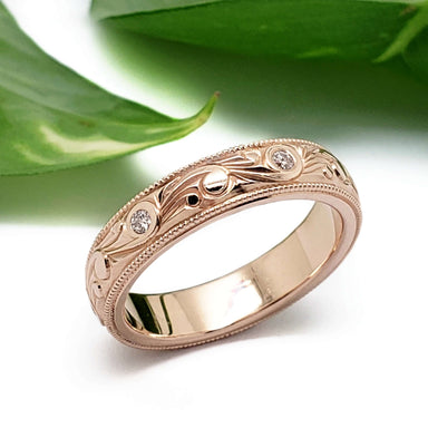 Handcrafted rose gold wedding ring with scroll engraving and diamonds, beside green leaves
