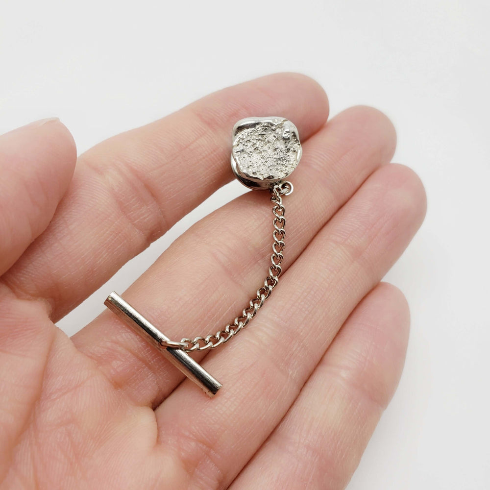 Hand holding sterling silver tie tack with concrete texture and chain, artisanal detail visible