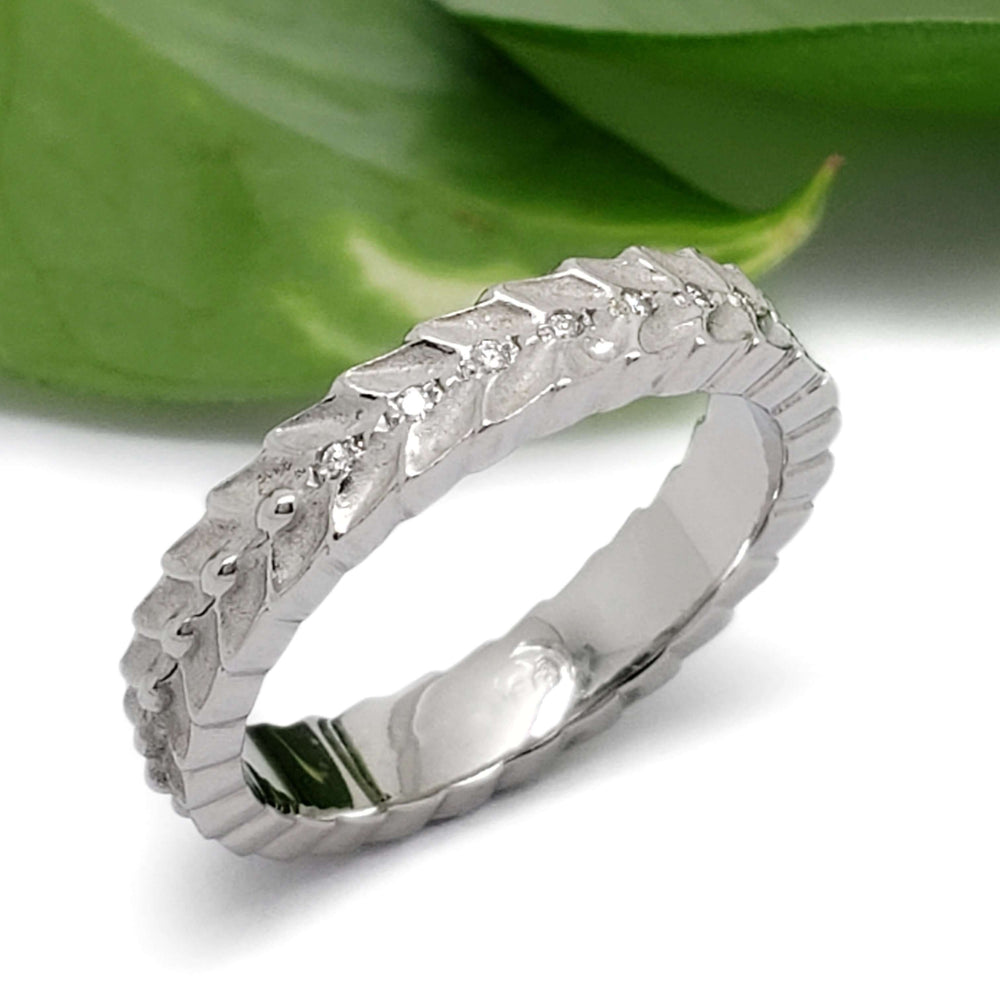 Handcrafted white gold leaf motif wedding band with diamonds, beside green leaf