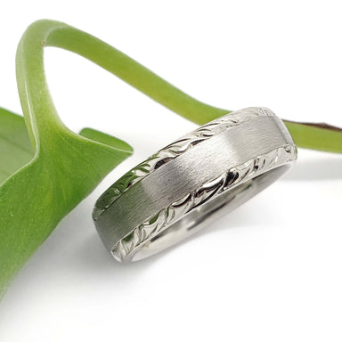 Handcrafted white gold wedding band with brushed finish and engraved edges beside a green leaf