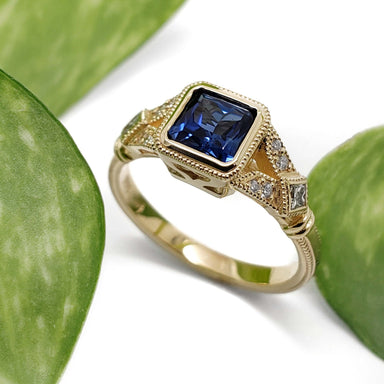 Handcrafted yellow gold engagement ring with blue sapphire and diamond accents, shown with leaves