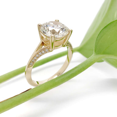 Handcrafted yellow gold engagement ring with round lab diamond and pave band beside green leaf