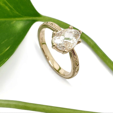 Handcrafted yellow gold pear lab diamond ring with engraved band beside a green leaf