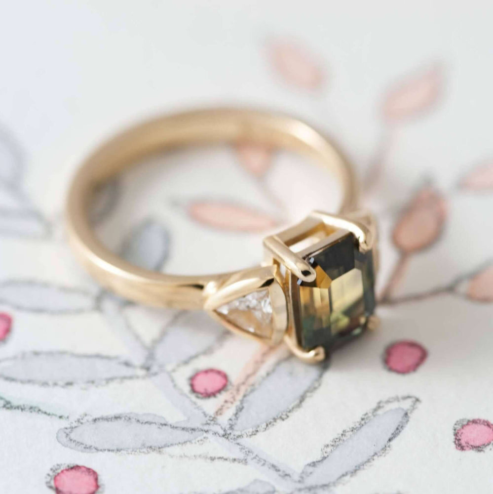 Handcrafted yellow gold ring with bi-coloured emerald cut sapphire and trillion diamonds on floral paper