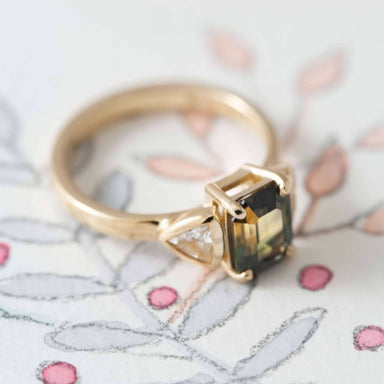 Handcrafted yellow gold ring with bi-coloured emerald cut sapphire and trillion diamonds on floral paper