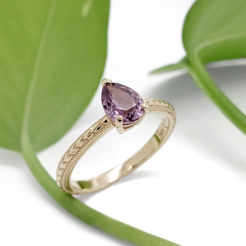 Handcrafted yellow gold ring with pink sapphire, engraved band, beside green leaves