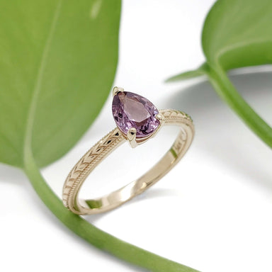 Handcrafted yellow gold ring with pink sapphire, engraved band, beside green leaves