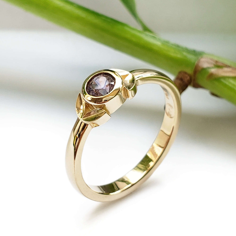Handcrafted yellow gold ring with purple Montana sapphire and leaf motif, shown near greenery