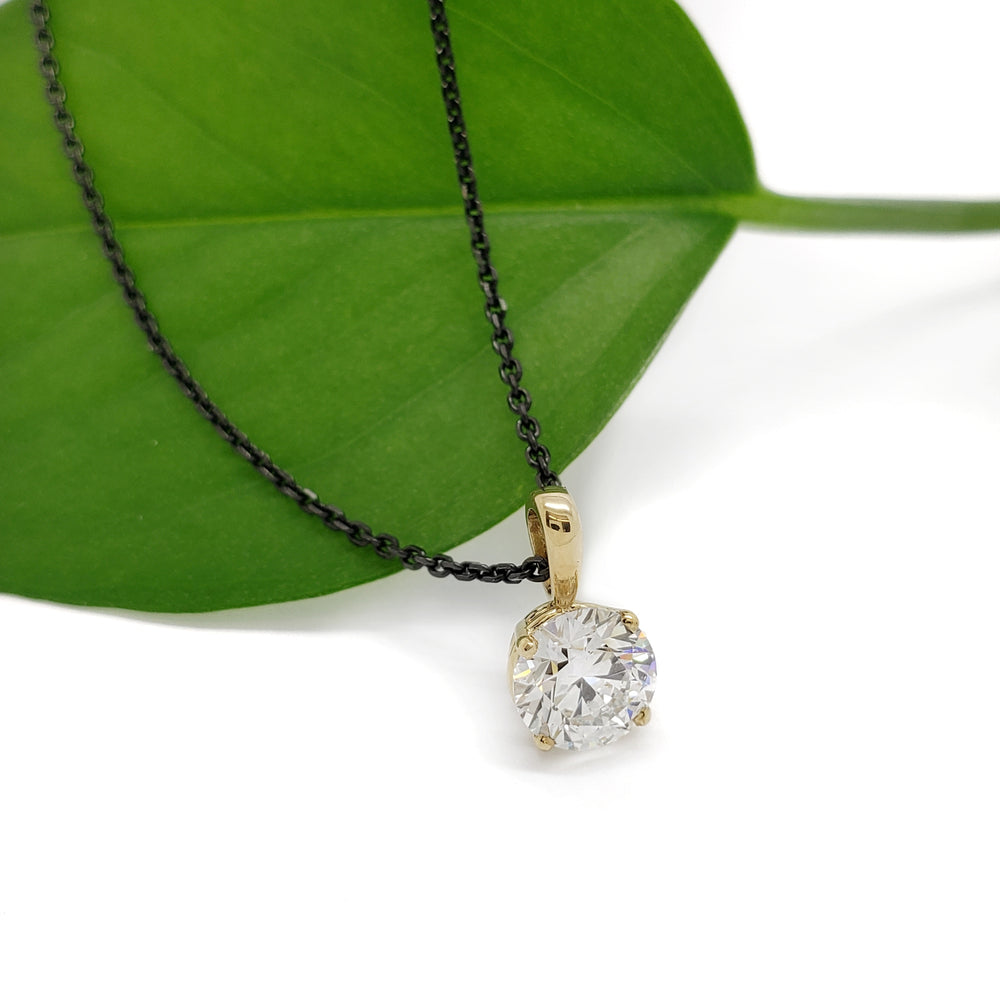A round-cut clear gemstone pendant on a delicate gold chain, styled on a vibrant green leaf against a clean white background.