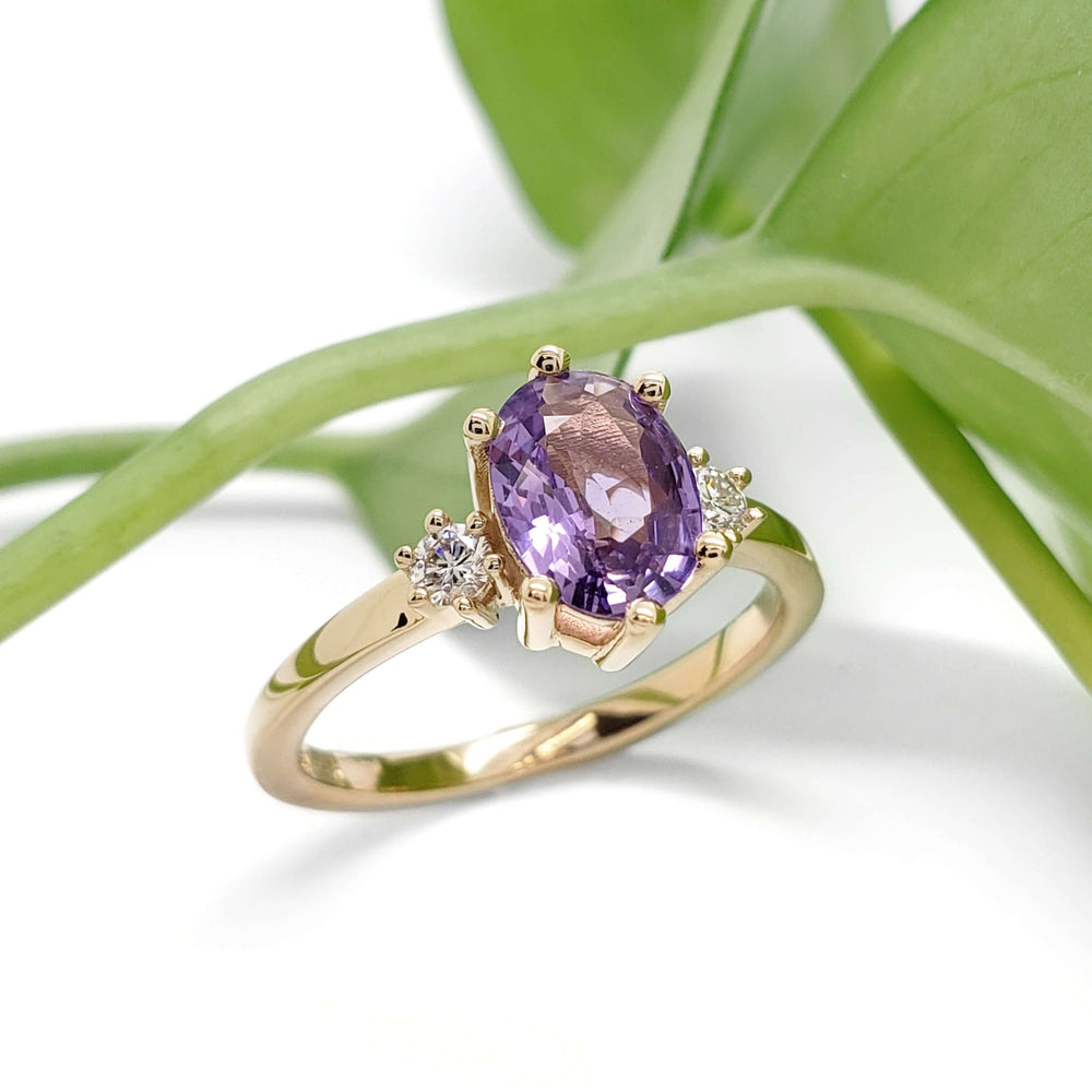 Lavender sapphire and lab diamond engagement ring in yellow gold, displayed with green leaves