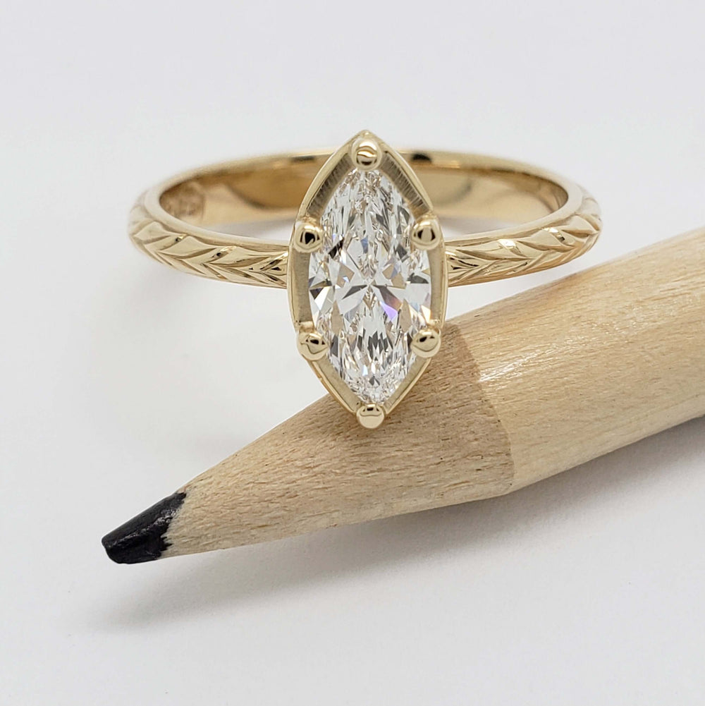 Marquise cut lab diamond engagement ring in yellow gold with engraved band, shown with pencil for scale