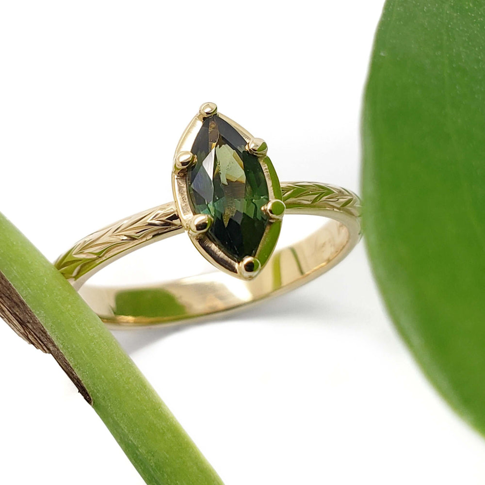 Marquise green sapphire ring in engraved 14kt yellow gold band beside green leaves