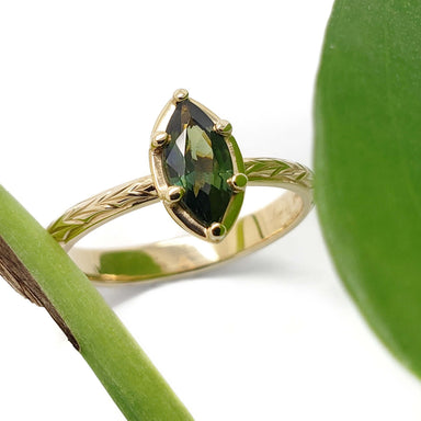 Marquise green sapphire ring in engraved 14kt yellow gold band beside green leaves