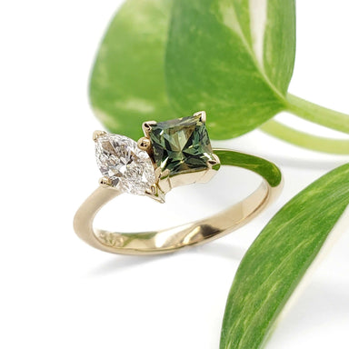 Moi et toi style yellow gold ring with diamond and teal sapphire beside green leaves