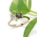 Moi et toi style yellow gold ring with diamond and teal sapphire beside green leaves