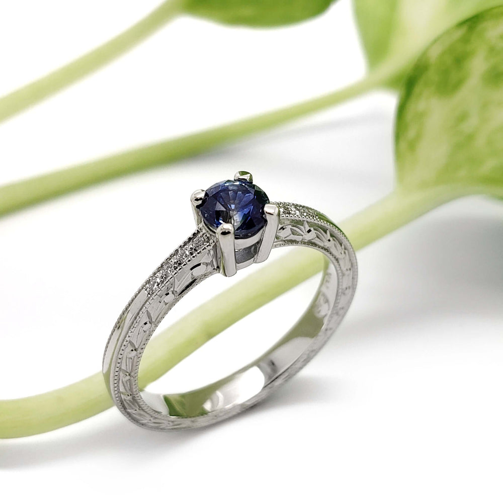 Montana sapphire and diamond engagement ring in white gold with engraved details, beside green plant