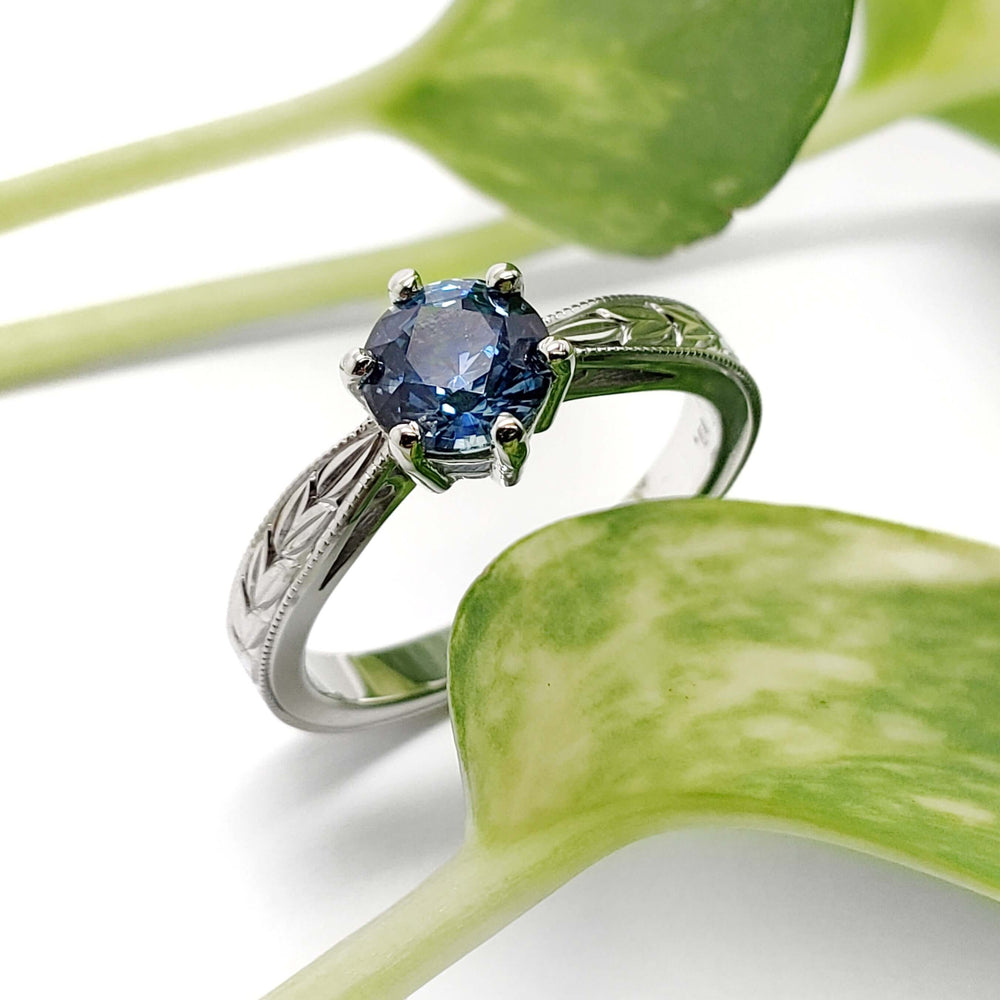 Handcrafted white gold Montana sapphire ring with engraved band, shown with green leaves