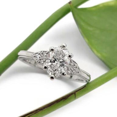 Oval lab diamond engagement ring in white gold with vintage-inspired accents, beside a green leaf