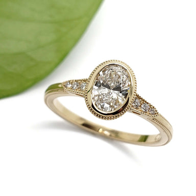 Oval lab diamond engagement ring in yellow gold with millgrain and pave details beside a leaf